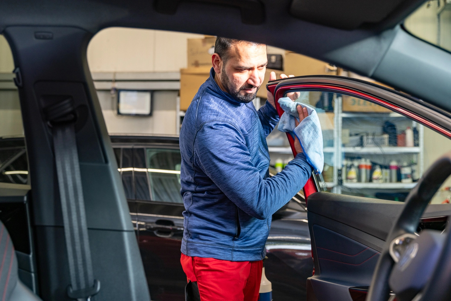 BlitzSauber das Reinigungszentrum Rellingen UG - Fahrzeugpflege - Mann in blauer Jacke und roter Hose reinigt in einer Garage die Innenseite einer roten Autotür mit einem blauen Tuch